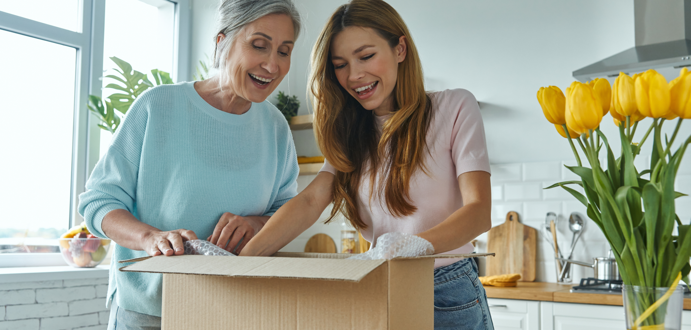 Two women smiling and opening a cardboard box in a bright kitchen, surrounded by yellow tulips. The scene conveys joy and anticipation.