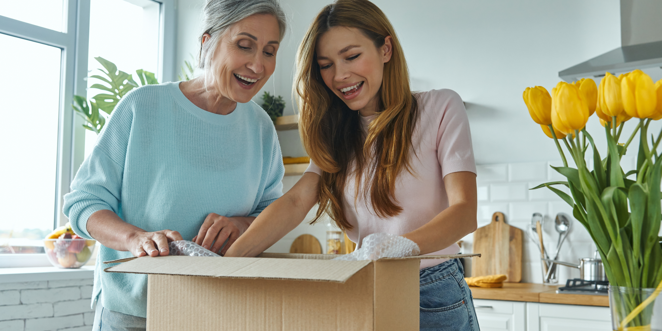 Two women smiling and opening a cardboard box in a bright kitchen, surrounded by yellow tulips. The scene conveys joy and anticipation.