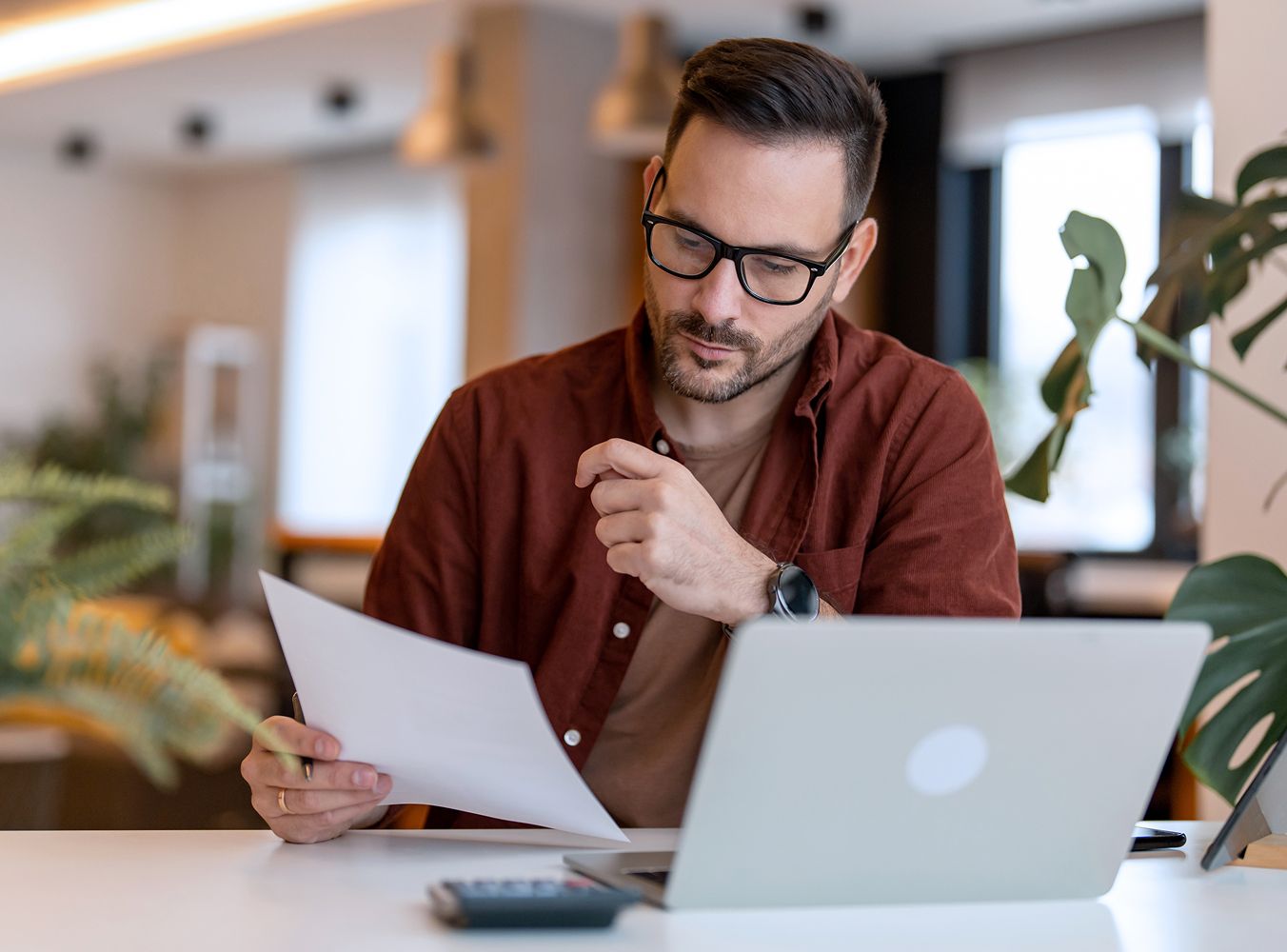 A man with glasses is thoughtfully reviewing a document at a desk with a laptop, plants and a calculator. The setting is a modern, well-lit office.