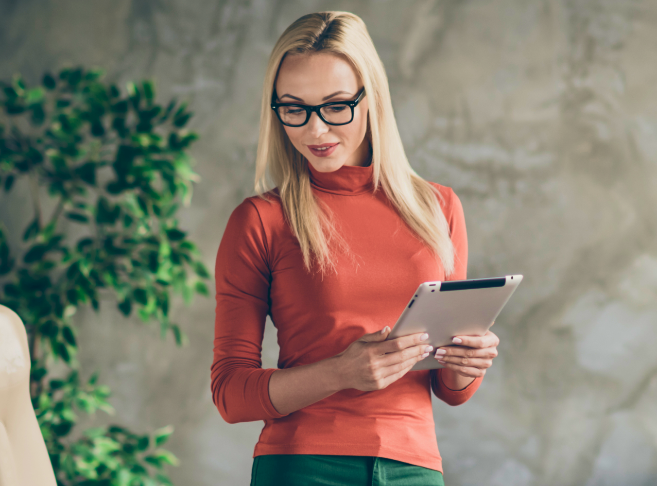 A woman with long blonde hair and glasses stands holding a tablet. She wears an orange turtleneck, exuding a relaxed, professional vibe.