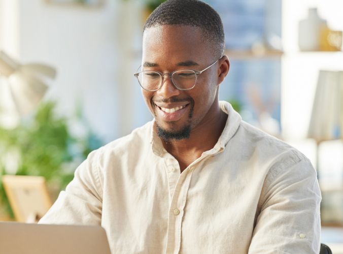 Smiling man with glasses using a laptop in a bright, plant-filled office. The scene conveys a positive and productive work atmosphere.