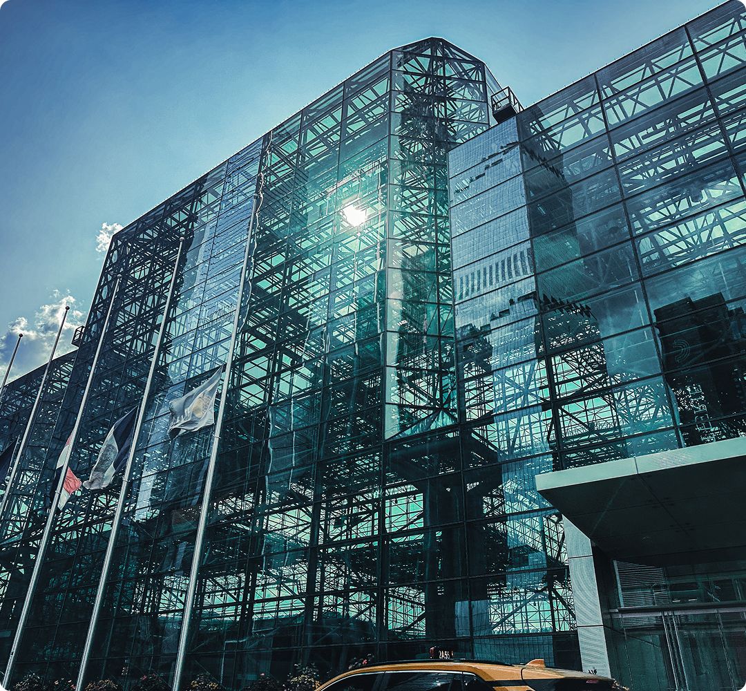 Glass facade of a modern building reflecting the sky, sun, and nearby flags. The structure has a geometric design, creating a sleek, futuristic vibe.