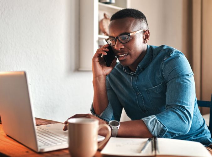A man in glasses and a denim shirt talks on a phone while working on a laptop. A notebook and coffee mug are on the table, creating a focused and productive scene.