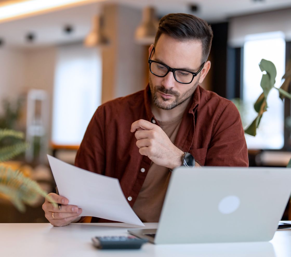 A man with glasses is thoughtfully reviewing a document at a desk with a laptop, plants and a calculator. The setting is a modern, well-lit office.