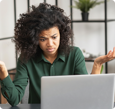 Woman in green shirt looks puzzled at laptop, raising hands in confusion. Background has shelves with a plant. Emotions convey frustration and bewilderment.