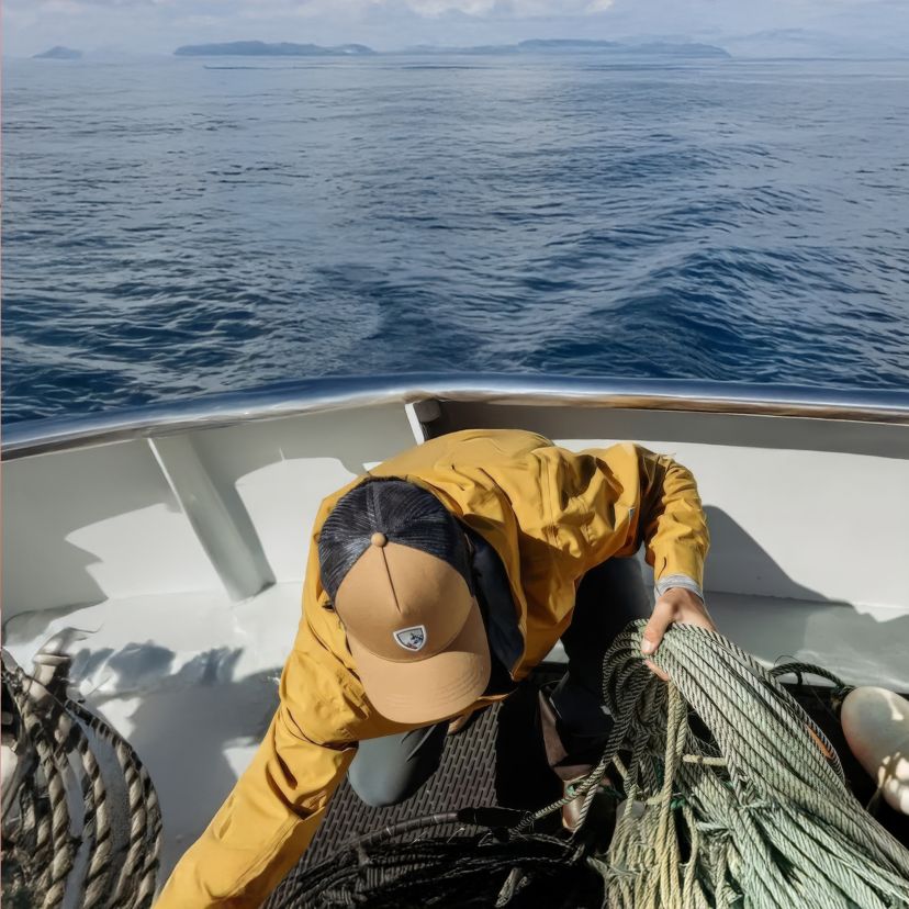 A person in a yellow jacket and cap organizes ropes on a boat deck against a backdrop of calm ocean and distant islands, conveying a sense of adventure.