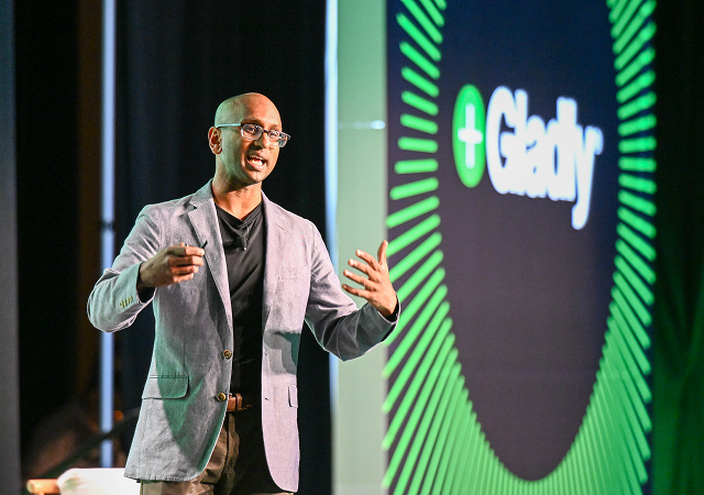 A man in a light gray blazer speaks energetically on stage with a "Gladly" logo displayed on a large screen. The setting is professional and engaging.