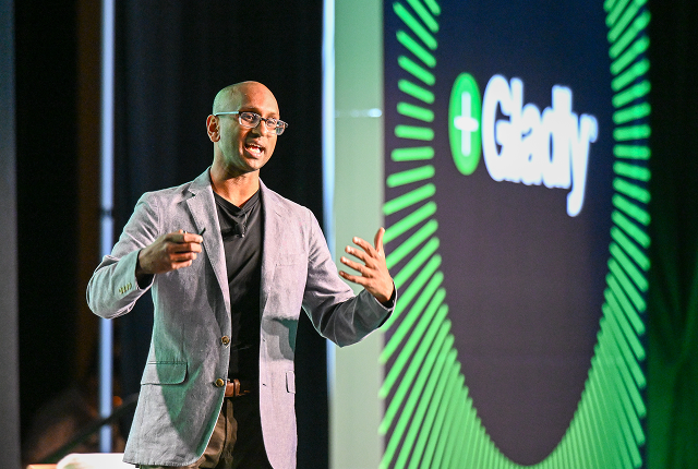 A man in a light gray blazer speaks energetically on stage with a "Gladly" logo displayed on a large screen. The setting is professional and engaging.