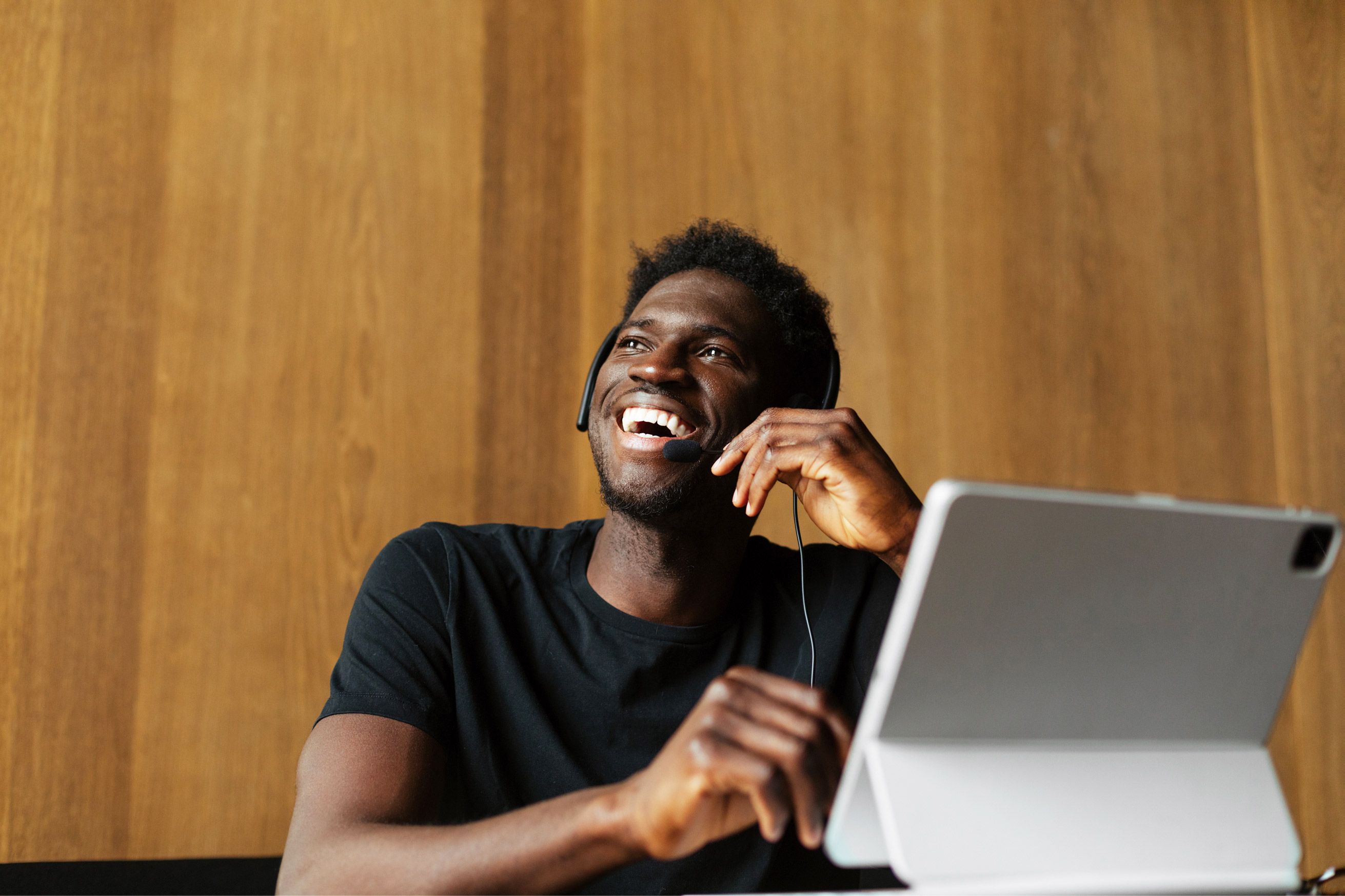 Smiling man with black hair and wearing headset, working on a laptop. He sits in front of a wooden wall. Relaxed atmosphere.