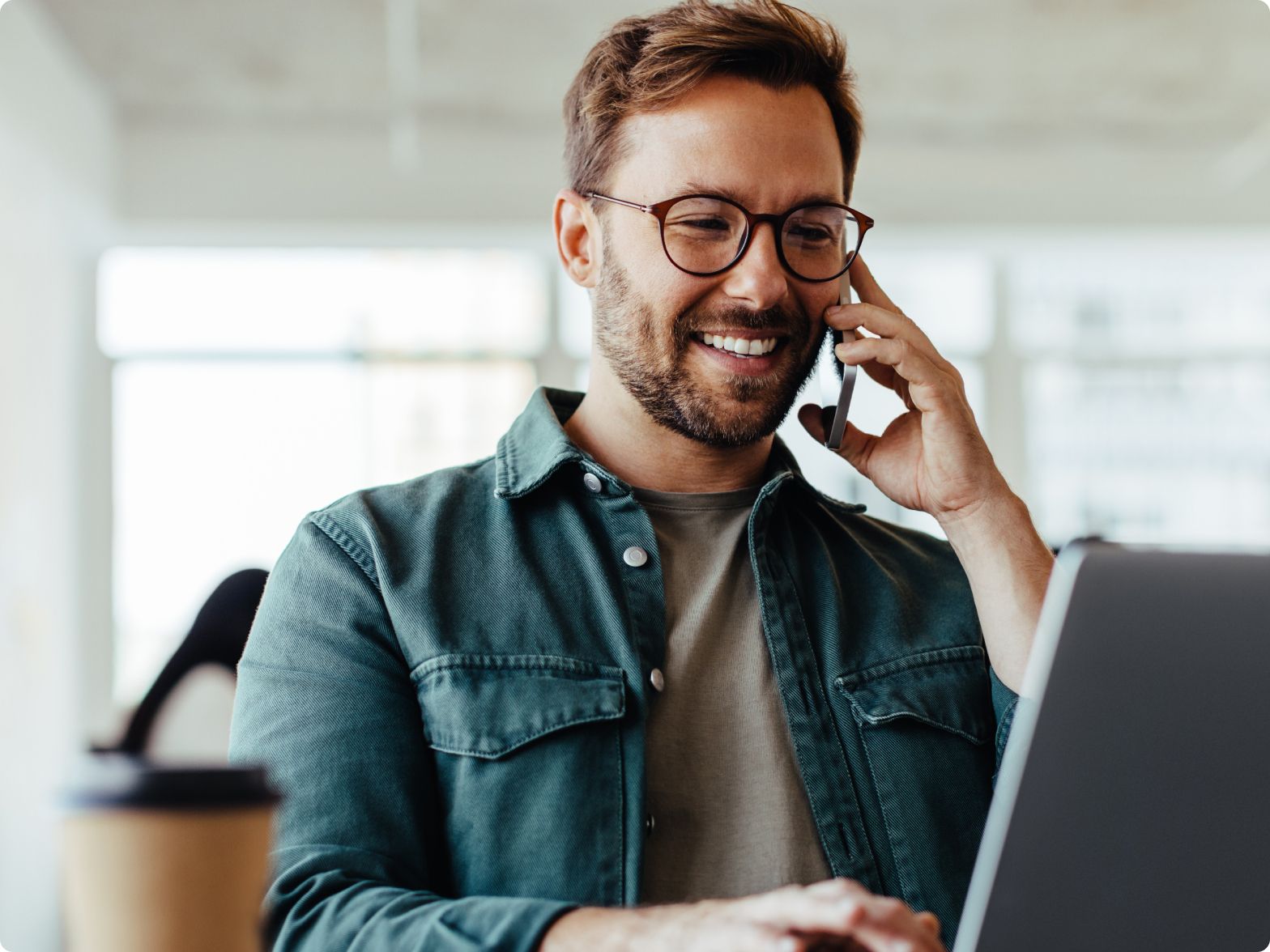 Smiling man with glasses uses a smartphone and laptop at a bright office desk. A takeaway coffee cup is in the foreground, conveying a productive atmosphere.