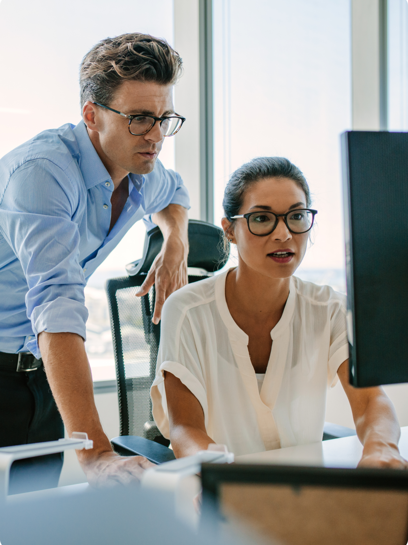 A man with glasses, wearing a green sweater, smiles while working on a computer in a bright office. A woman in a striped shirt works behind him.