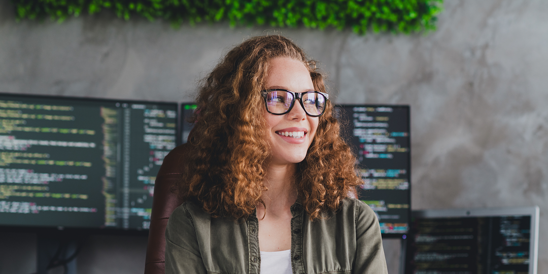 Smiling woman with curly hair and glasses sits in front of computer monitors displaying code. The atmosphere is focused yet relaxed, with a green plant above.