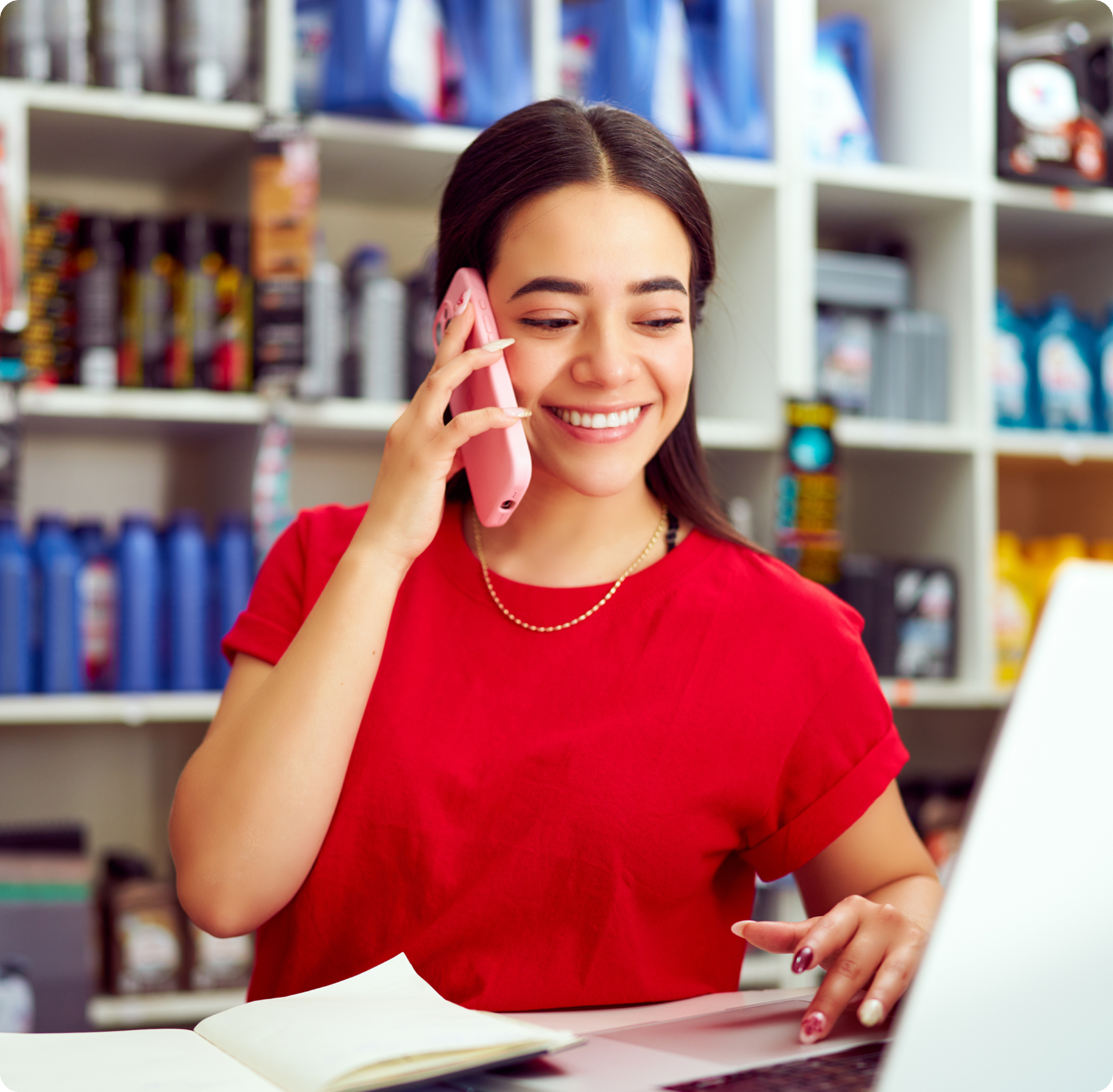 Smiling woman in a red shirt talks on a pink phone while using a laptop in a store. Shelves with various products are visible in the background.