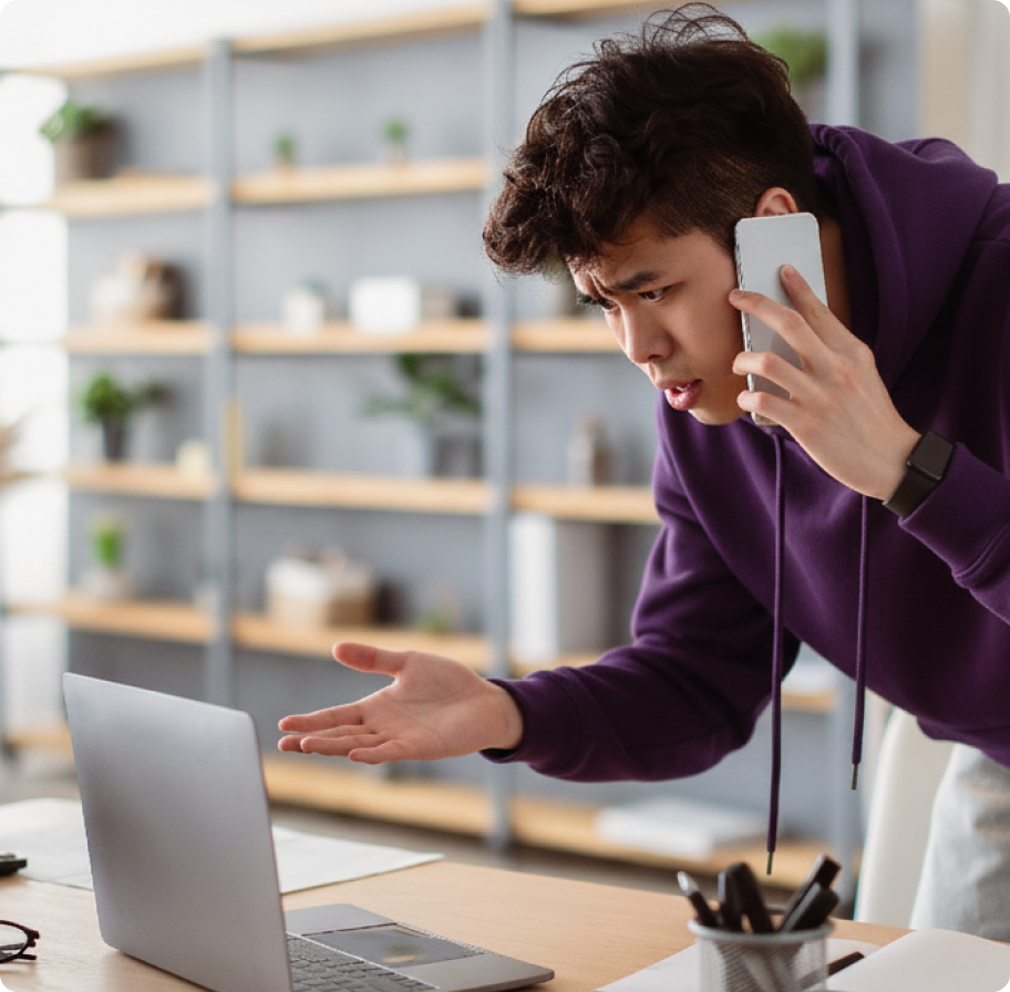 A man in a purple hoodie, looking frustrated, holds a smartphone to his ear while gesturing at a laptop. The setting is a modern home with shelves.