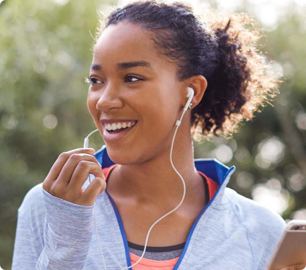 A smiling woman enjoys music through earphones outdoors. Wearing a blue jacket, she holds a phone, exuding joy and warmth in a sunlit setting.