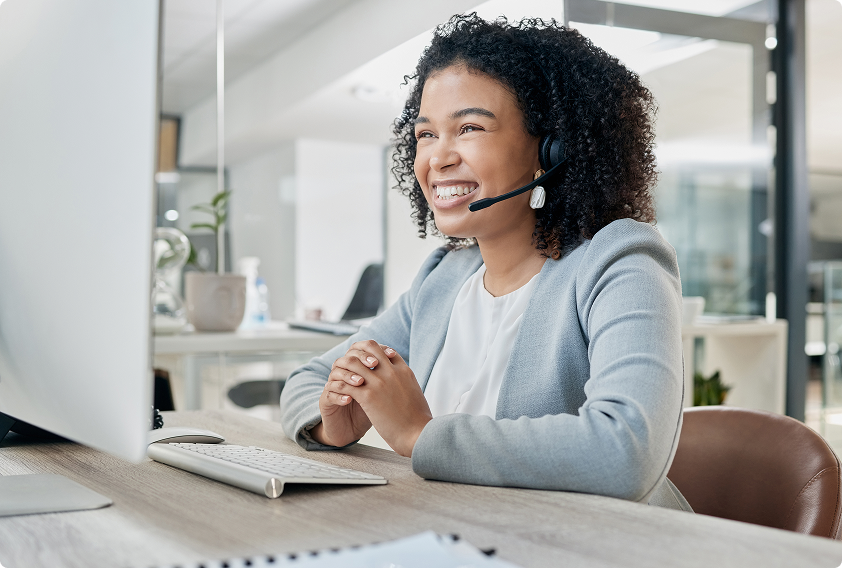 Smiling woman with headphones sits at a desk in a modern office, engaging with a computer. She wears a light blazer, conveying professionalism and warmth.