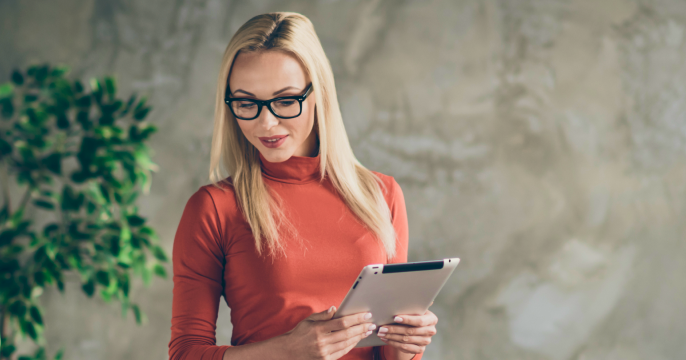 A woman with long blonde hair and glasses stands holding a tablet. She wears an orange turtleneck, exuding a relaxed, professional vibe.