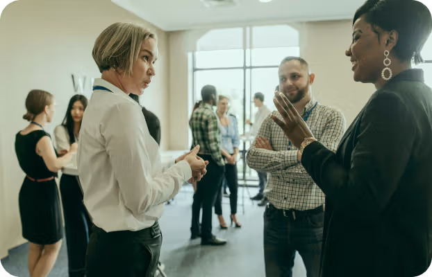A group of professionals engaged in conversations at a networking event. Three people are in focus, discussing animatedly, with more interactions in the background. The atmosphere is lively and collaborative.
