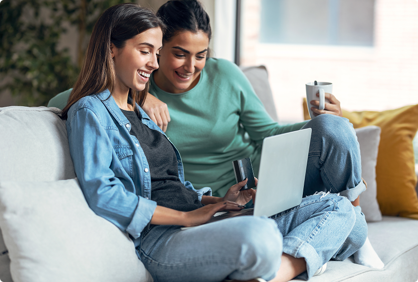 Two women sitting on a sofa, smiling at a laptop. One holds a credit card; the other has a mug, conveying a cozy, relaxed online shopping moment.