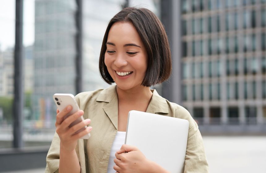 Lady outside holding her laptop in one hand and smiling at her phone on the other