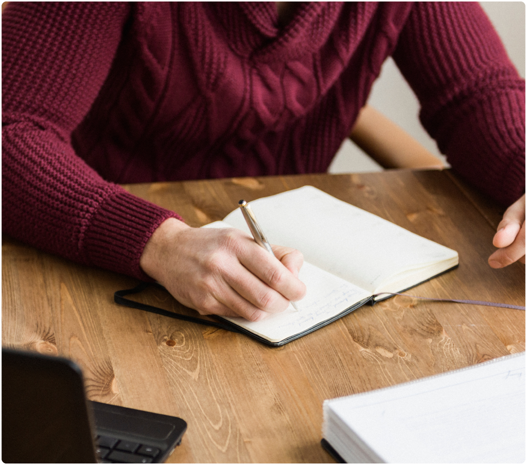 A person in a maroon sweater writes in a notebook with a pen at a wooden desk. A laptop and a notebook are on the desk, suggesting a study or work setting.