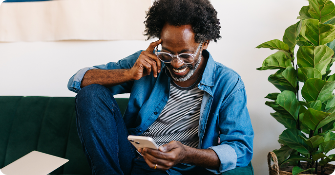A man with curly hair and glasses smiles while looking at his phone. He's wearing a denim shirt and striped shirt, sitting on a green couch.