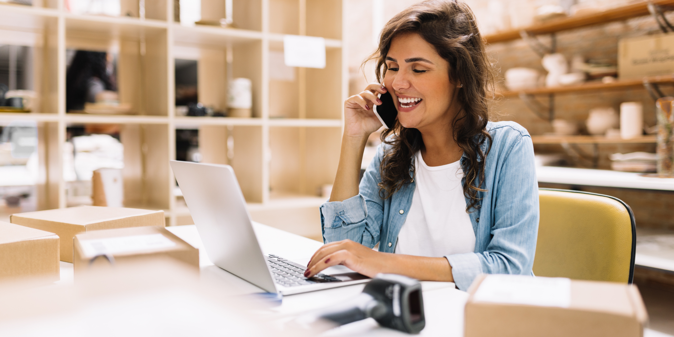 Woman smiling while talking on the phone in an office. She sits at a desk with a laptop and several cardboard boxes, suggesting a business or e-commerce setting.