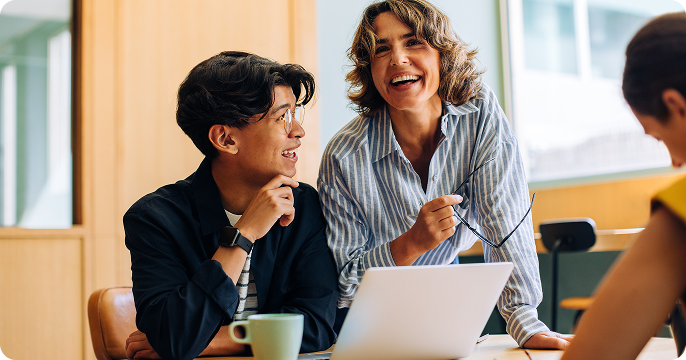 A cheerful woman and a young adult sit at a table with a laptop and coffee mug. They are laughing and appear to be having an engaging conversation.
