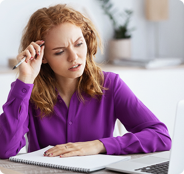 A woman with red hair wearing a purple shirt looks confused as she writes in a notebook at a desk, pen in hand, in a bright, modern workspace.