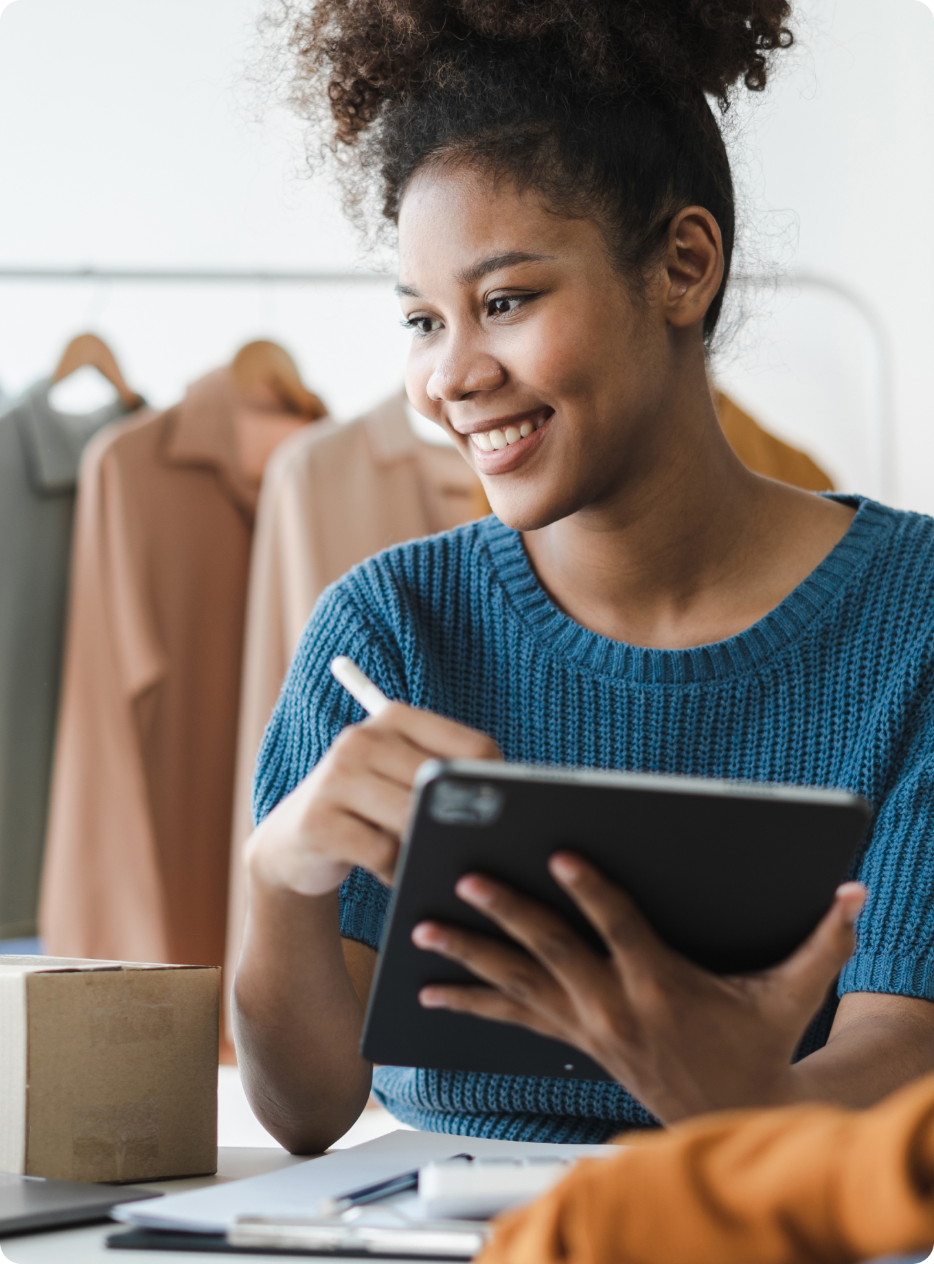 Smiling woman in a blue sweater works on a tablet with a stylus. Background shows clothes on hangers, suggesting a creative or retail setting.