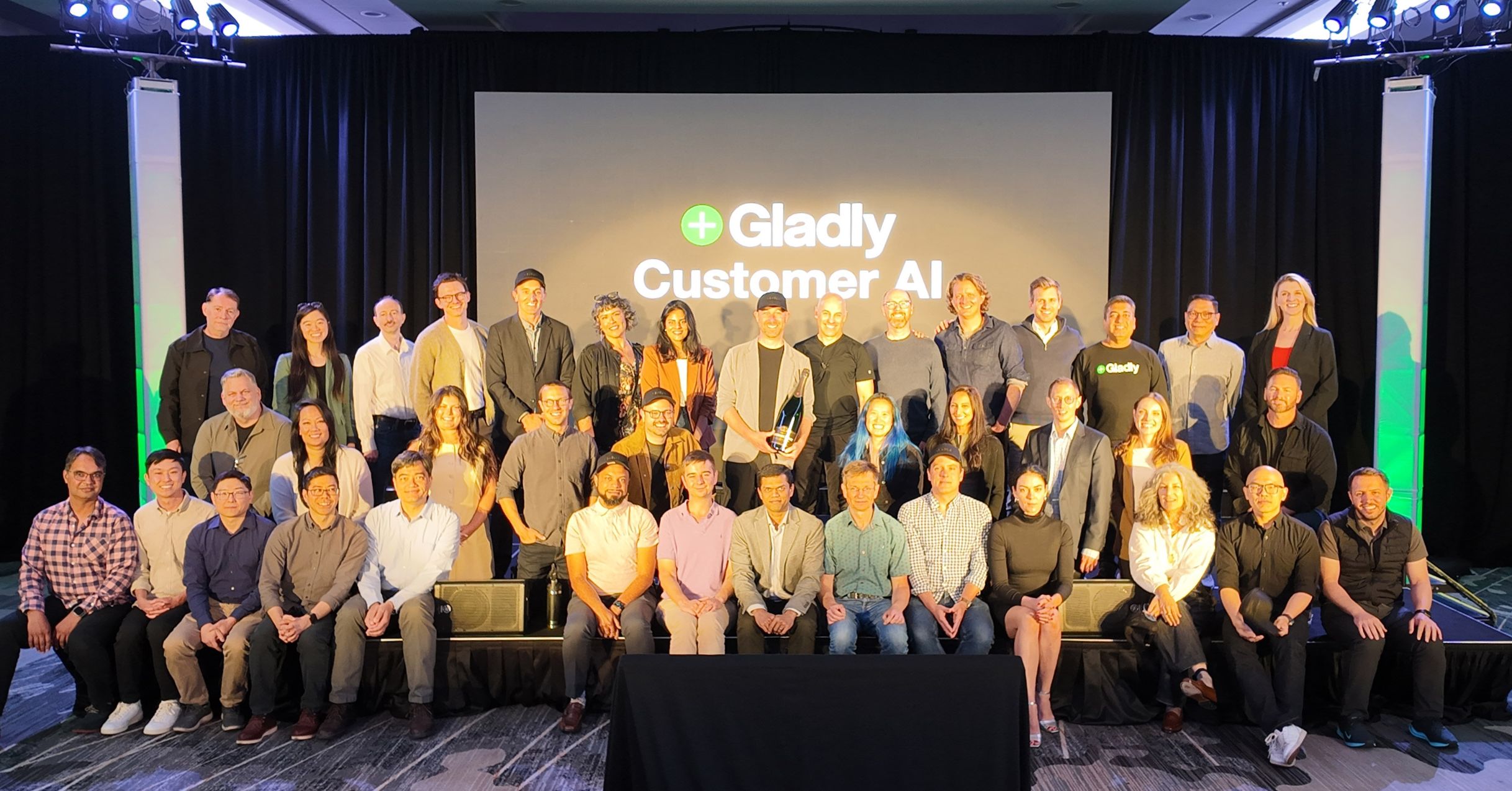 A diverse group of around 40 people pose on stage in front of a screen displaying "Gladly Customer AI." They appear happy and professional.