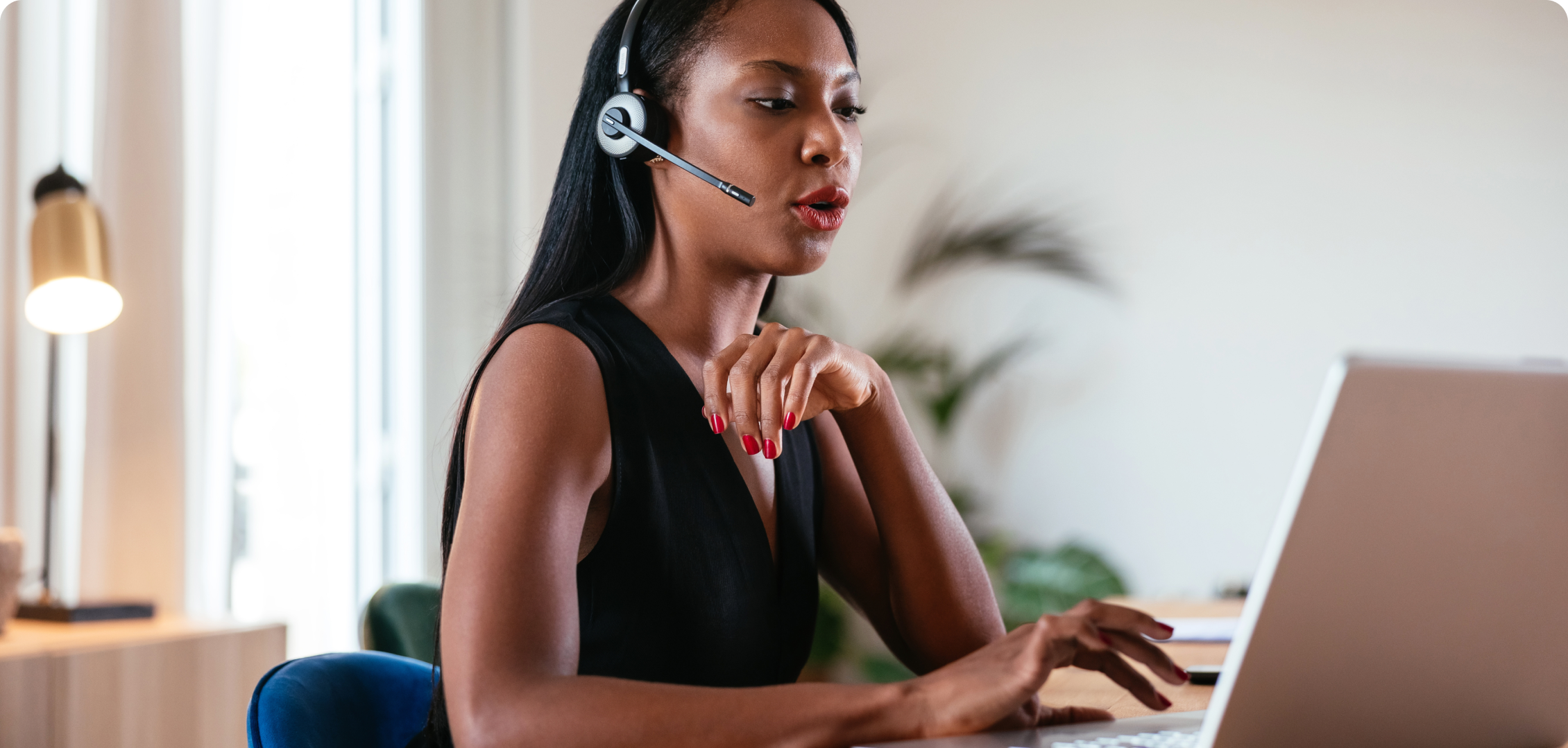 A woman in a black top works at a laptop, wearing a headset with a microphone. The setting is a well-lit, modern home office, conveying focus and professionalism.