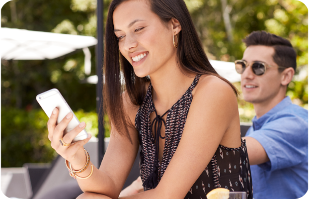 A smiling woman sits outdoors, looking at her smartphone, with a man in sunglasses in the background. The scene is relaxed and sunny, suggesting leisure.