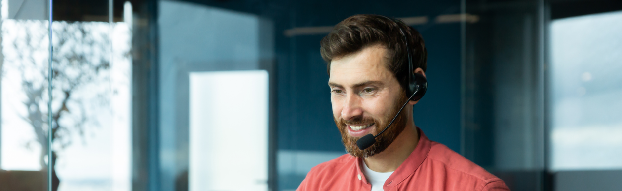 Bearded man wearing a phone headset and a salmon-colored shirt gesturing at a laptop