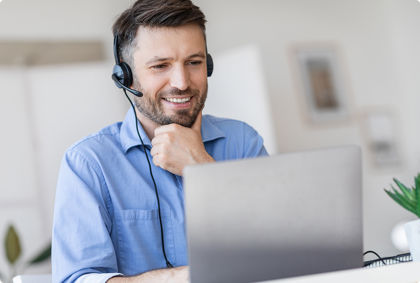 A man in a blue shirt wearing a headset smiles while video conferencing on a laptop in a bright, modern home office. A small plant is visible beside him.