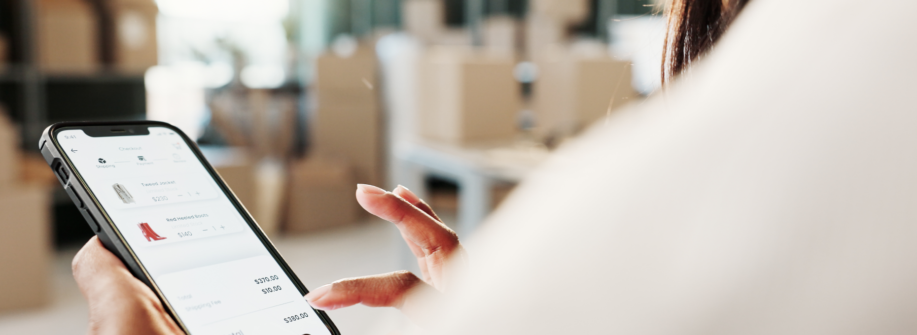 Close-up over-the-shoulder view of someone reviewing a checkout screen on their phone, with an order summary and a "confirm order" button visible, standing in a warehouse with stacked shipping boxes in the background.