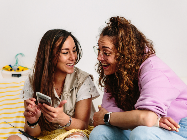 Two Gen Z women sit together looking at a smartphone and smiling while shopping online