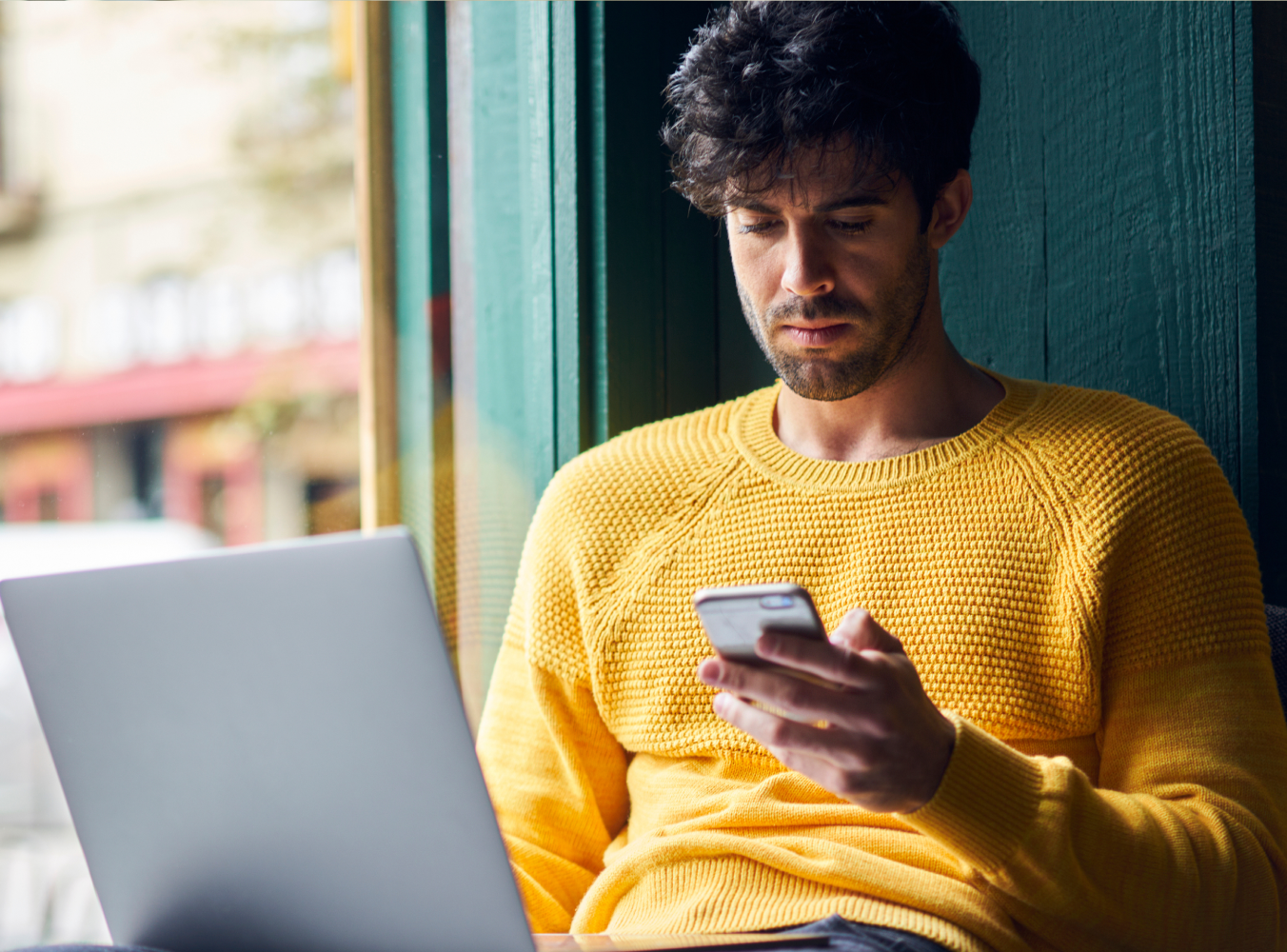 A man in a yellow sweater sits by a window, focused on his smartphone. He has a laptop on his lap and a coffee cup beside him, conveying a relaxed, studious mood.