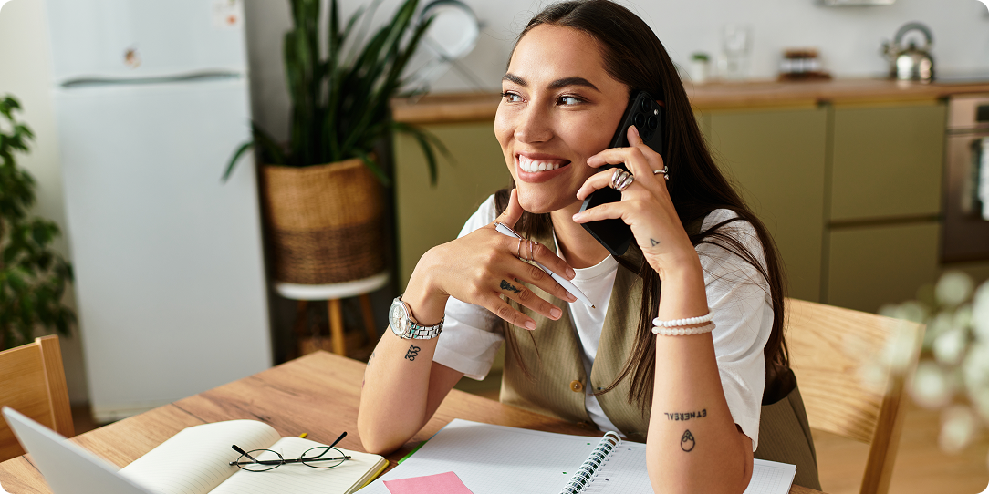 A woman smiling while talking on the phone sits at a table with an open notebook. She appears relaxed in a bright kitchen, exuding a cheerful mood.