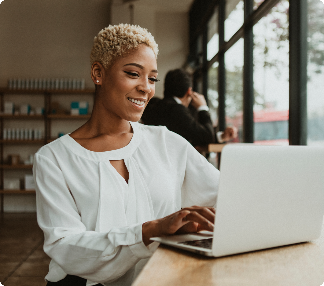 A woman in a white blouse types on a laptop, smiling warmly. She sits by large windows in a bright cafe, creating a cheerful and focused atmosphere.