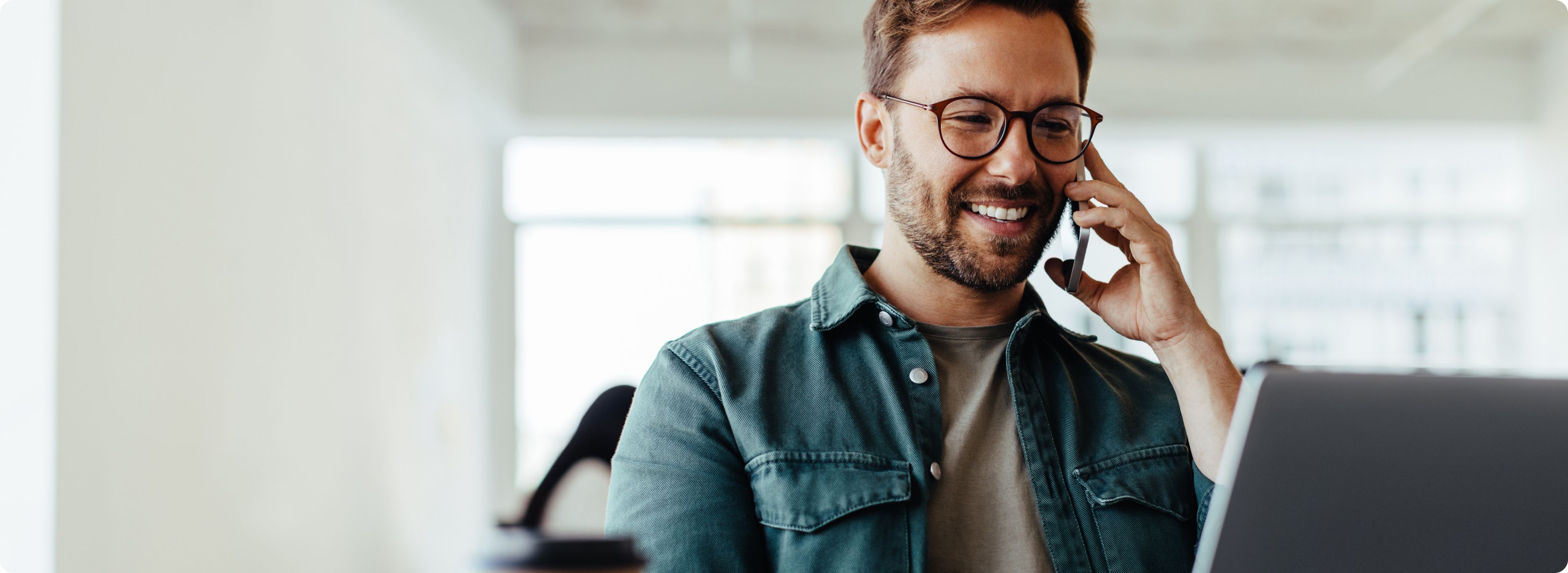 Smiling man with glasses uses a smartphone and laptop at a bright office desk. A takeaway coffee cup is in the foreground, conveying a productive atmosphere.