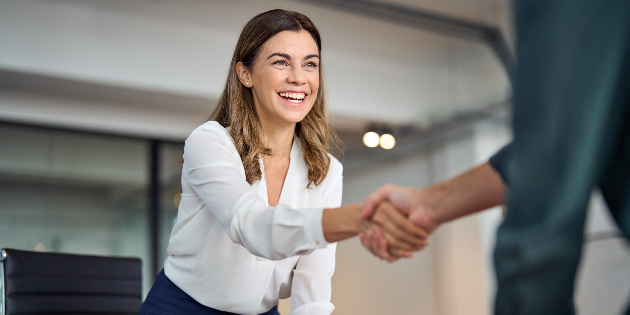 A woman in a white blouse smiles warmly while shaking hands with someone off-camera in an office setting, conveying friendliness and professionalism.