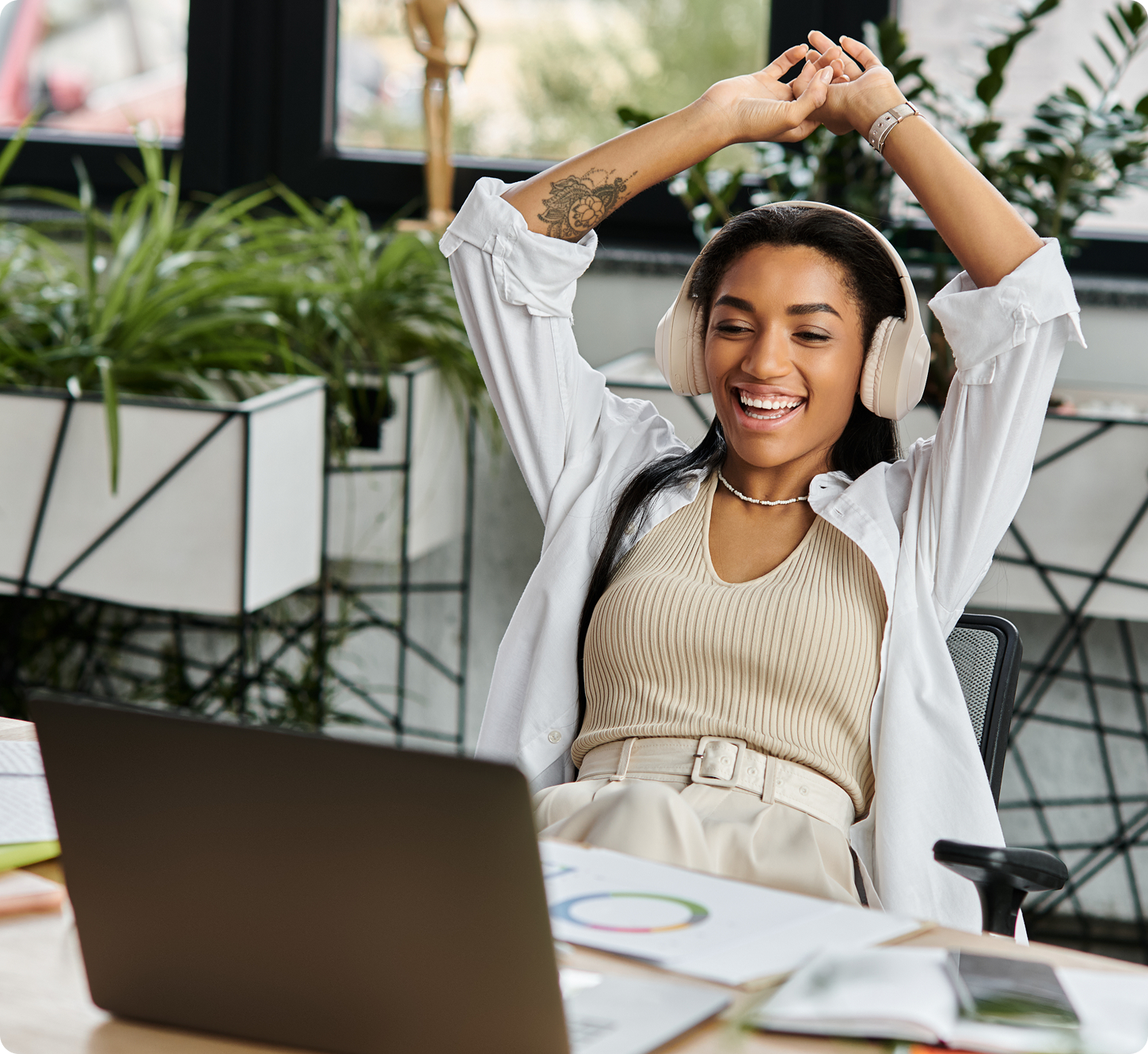 Young woman sitting at a desk, wearing headphones, and stretching with a joyful expression. Relaxed office setting with plants around her.