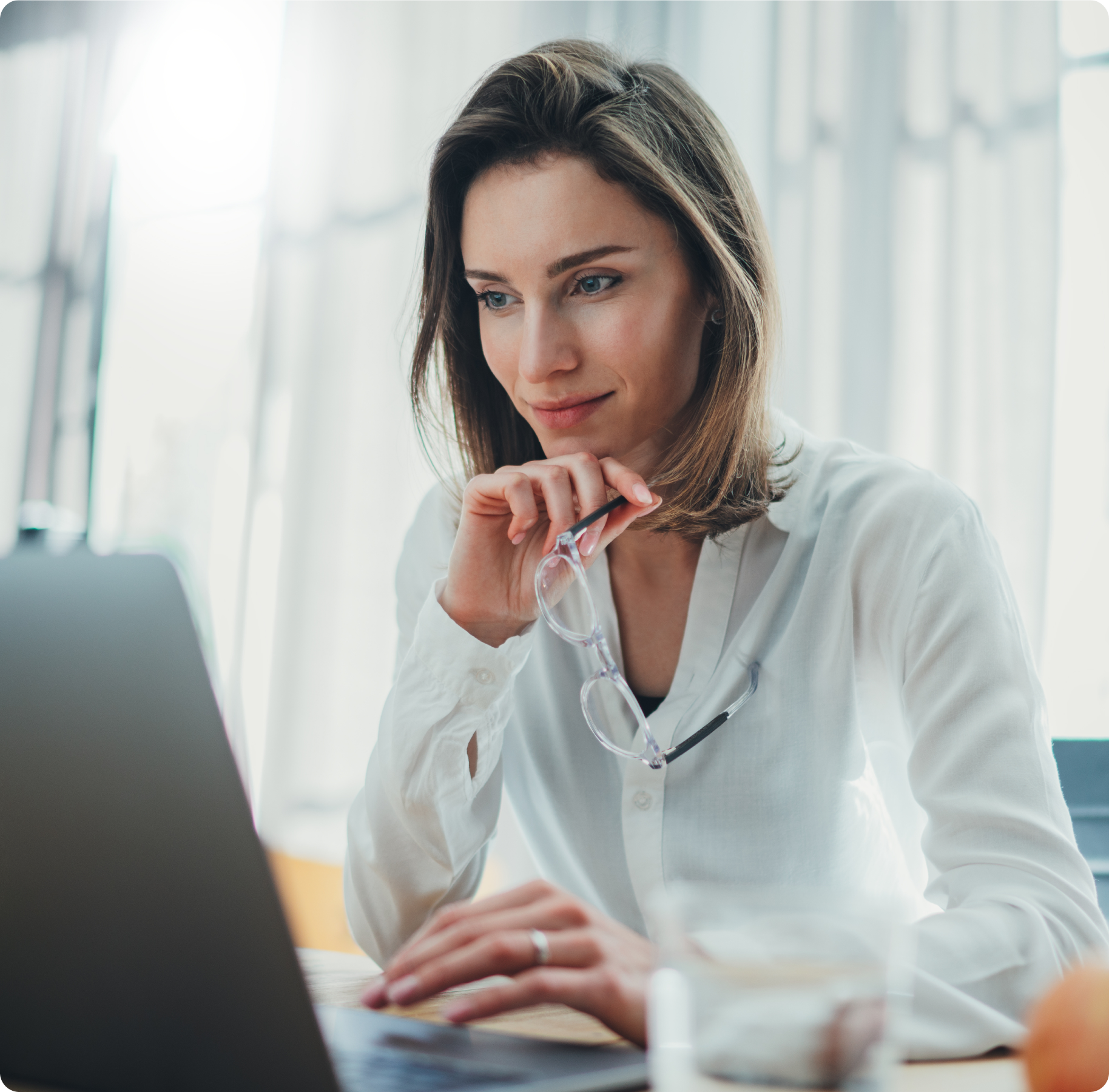 A woman in a white blouse sits at a desk, gazing thoughtfully at a laptop while holding eyeglasses. The scene is bright and focused, suggesting concentration.