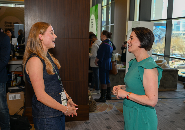 Two women stand in a well-lit conference hall, engaged in a friendly conversation. Both are smiling warmly, creating an atmosphere of connection and positivity.
