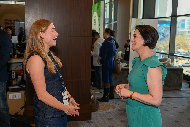 Two women stand in a well-lit conference hall, engaged in a friendly conversation. Both are smiling warmly, creating an atmosphere of connection and positivity.