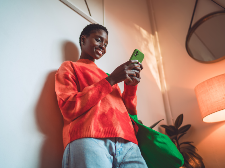 Smiling woman in an orange sweater leans against a wall at home, shopping on her phone