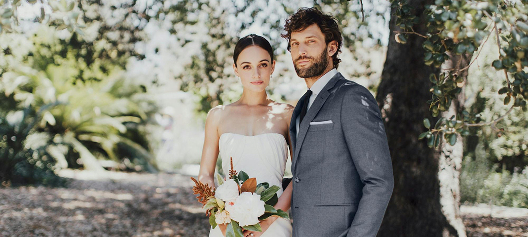 A bride in a strapless white dress holds a bouquet, standing beside a groom in a gray suit and tie. They are under a tree, exuding a serene and elegant vibe.