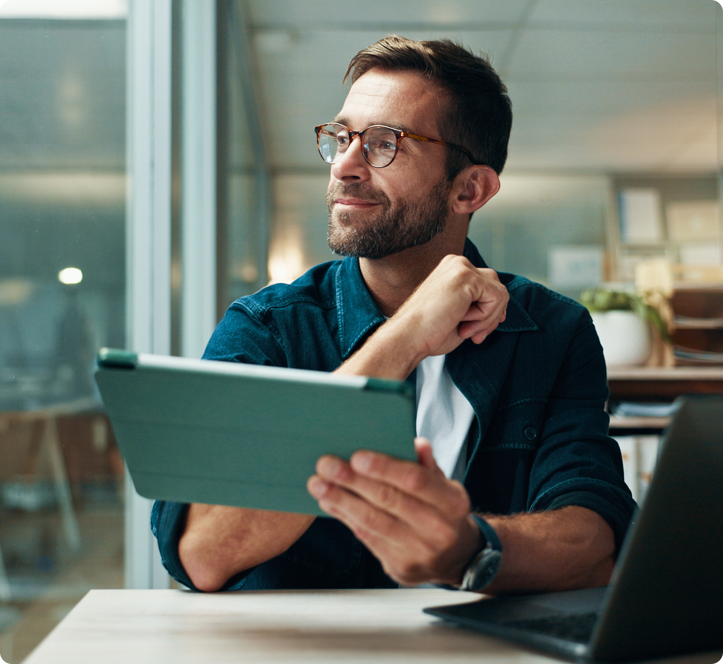 Smiling man with glasses using a laptop in a bright, plant-filled office. The scene conveys a positive and productive work atmosphere.