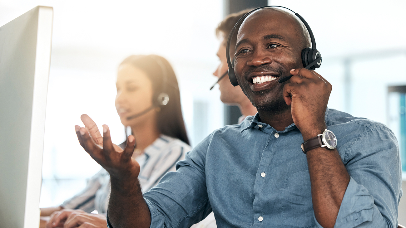 Smiling call center representative wearing a headset and a blue shirt, gesturing while talking. Two colleagues with headsets work in the background. Bright, professional office setting.